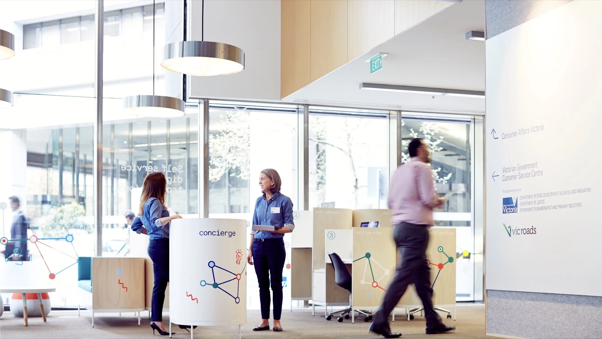 A bright Victorian Government service centre with concierge staff helping visitors at a branded kiosk.